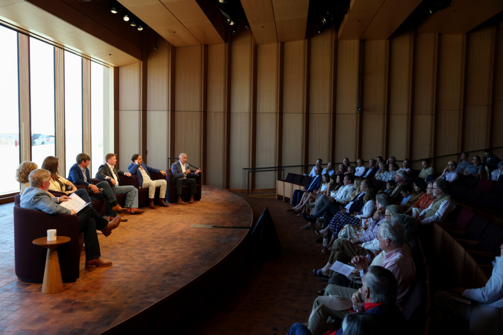 Panelists speak to a full audience in the wood-paneled auditorium of the Theodore Roosevelt Presidential Library, seated on a curved stage beside tall windows overlooking the Badlands.