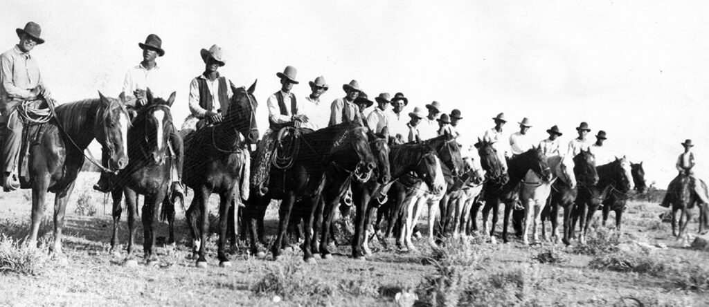 Vintage black-and-white photo of a long line of cowboys on horseback, wearing hats and Western wear, posed in a diagonal row across an open plain.