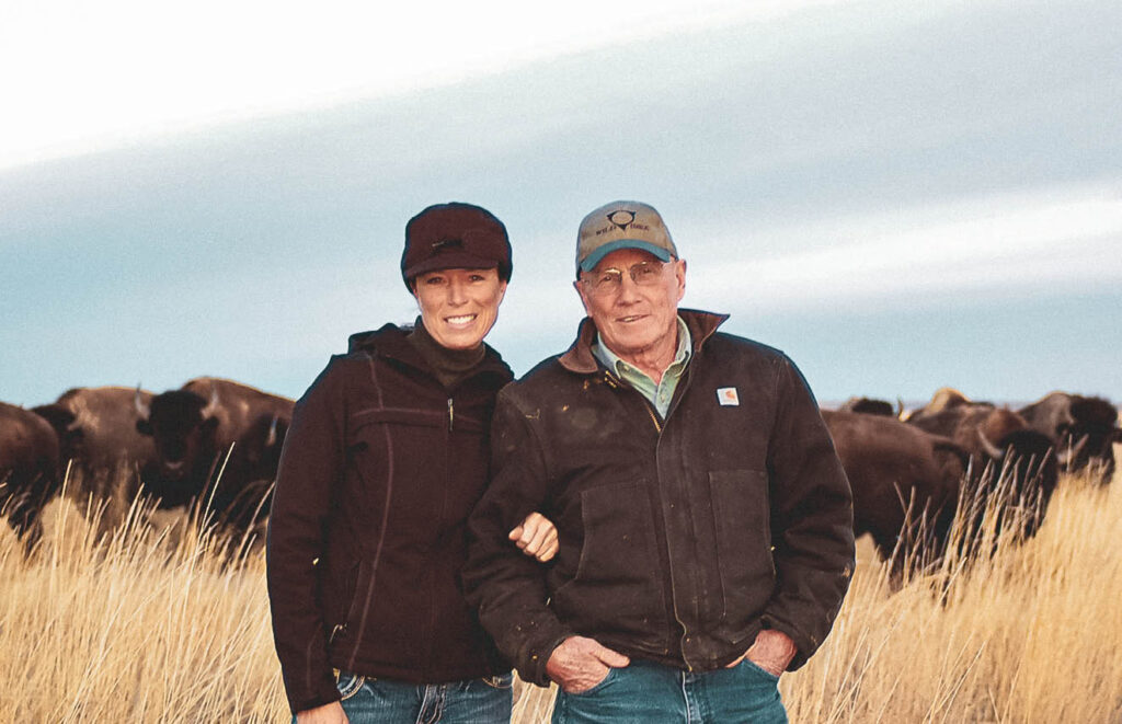 A woman and an older man in caps and jackets stand arm in arm smiling in front of a bison herd grazing in tall golden grass under a hazy sky.