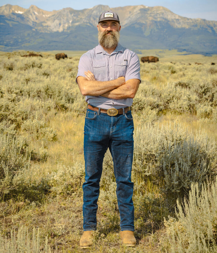 A bearded man in a cap and denim, arms crossed, stands in a sagebrush field wearing a belt buckle with a bison emblem, with distant bison and mountains behind him.